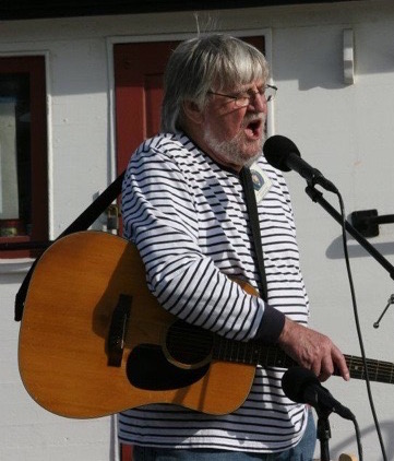 Image of Dick in a striped shirt standing up and singing into a microphone. He is wearing a guitar.