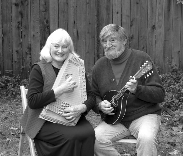Black and white image of a white woman and man sitting down and looking at the camera and smiling. They are both holding musical instruments.
